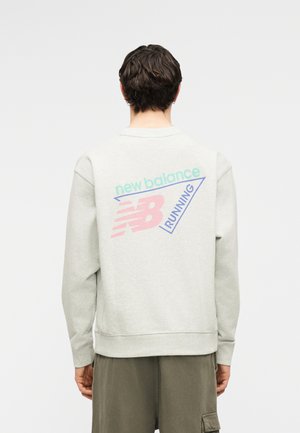 Person sett bakfra iført en lys grå genser med pastellfarget "new balance RUNNING"-logo og olivengrønne cargobukser.