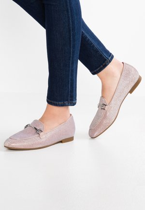Person iført glitrende lyserøde loafers med metal spænde, kombineret med mørkeblå skinny jeans mod en hvid baggrund.