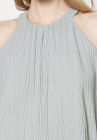 Vestido sin mangas en un gris claro con un escote alto, que presenta una tela texturizada y plisada, y una tira trasera lisa y redondeada.