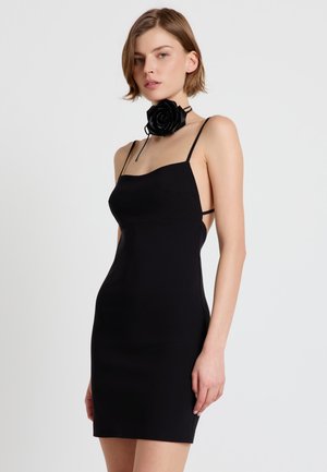 Donna che indossa un abito nero senza maniche aderente con spalline sottili e una grande collana choker a forma di rosa nera, in piedi davanti a uno sfondo semplice.