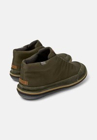 Olive green shoes with a textured fabric upper and black soles, featuring a slight heel and a branded tag on the back.