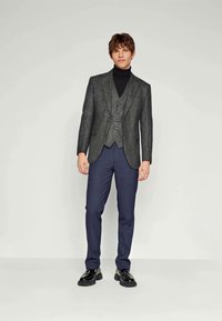 Traje de tres piezas gris de tela con solapa de muesca, cuello vuelto negro, pantalones azul marino y zapatos negros. Textura suave, corte entallado.