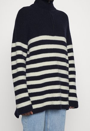 Personne portant un pull zippé rayé bleu marine et blanc à manches longues et un jean bleu clair, visible du cou aux hanches.