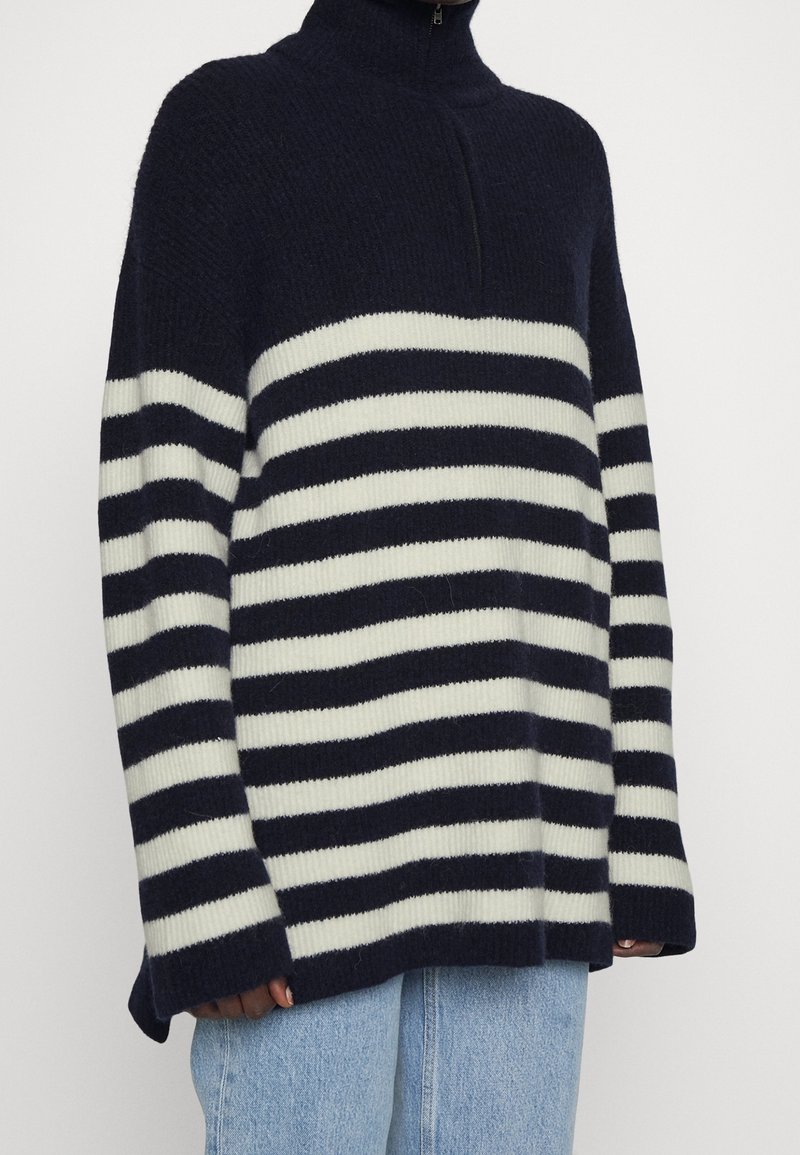Personne portant un pull zippé rayé bleu marine et blanc à manches longues et un jean bleu clair, visible du cou aux hanches.