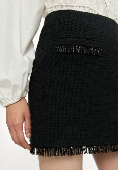 Jupe mini en tweed noir avec une surface texturée, une poche unique et des détails en frange noire au niveau de l'ourlet. Associée à une blouse blanche.