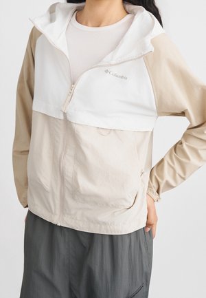 Persoon draagt een beige-witte Columbia-jas met rits, staat met één hand op de heup, gecombineerd met donkergrijze broek.
