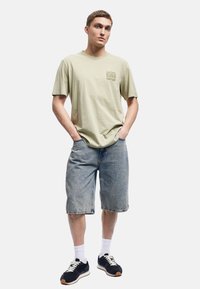 Ung mand iført en beige grafisk t-shirt, knæ-lange denim shorts, hvide sokker og marineblå sneakers, stående med hænderne i lommerne.