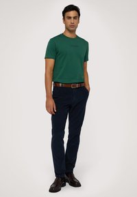 T-shirt in cotone verde con maniche corte e scollo rotondo, abbinato a pantaloni blu scuro e scarpe di pelle marrone. Una cintura marrone evidenzia la vita.