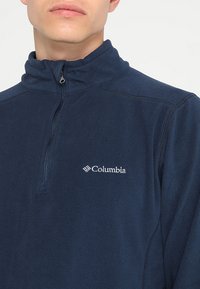 Mornarsko moder flis pulover s visokim ovratnikom in četrtno zadrgo, ki ima bele Columbia logo na levem prsnem delu in teksturirano tkanino.