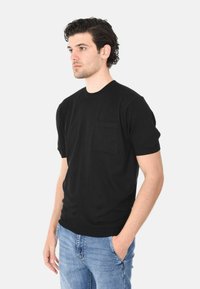 Maglione nero a maniche corte con scollatura rotonda e tasca sul petto. Materiale in maglia morbido; design aderente. Indossato con jeans di denim blu.