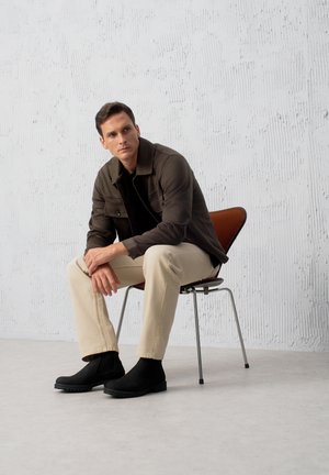 Siedzący męski model w brązowej kurtce na czarnej koszuli, w beżowych spodniach i czarnych butach, na krześle w stylu mid-century, przy fakturowanej ścianie.