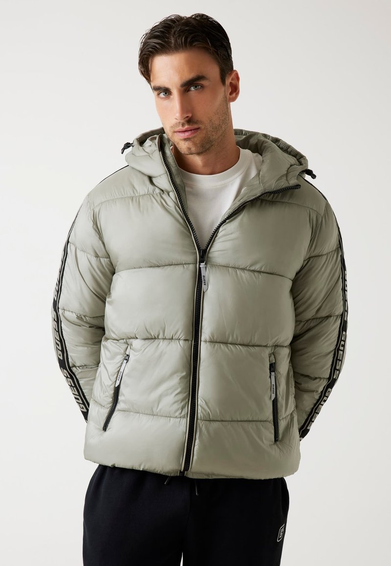 Giacca a vento verde chiaro con zip frontale, cappuccio e tasche laterali. Presenta strisce nere sui lati con testo del logo. Texture liscia e lucida.