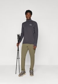 Szürke hosszú ujjú póló magas gallérral, olívazöld nadrág és szürke túracipő. Fekete túrabottal a kezében, részletes hardver díszítéssel.