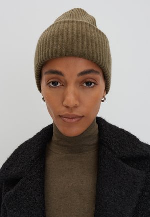 Olive green ribbed knit beanie with a pointed top, worn over a dark textured coat and an olive turtleneck sweater, creating a layered look.