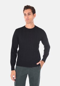 Uomo con capelli ricci corti, che indossa un maglione nero a treccia e pantaloni verde scuro, in piedi con una mano in tasca.