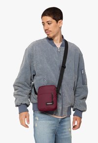 Borsa a tracolla in nylon bordeaux con tasca con zip, tracolla regolabile e patch logo, indossata sopra una giacca bomber blu.