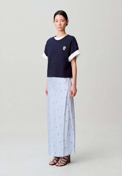 T-shirt corta blu navy con maniche arrotolate bianche, sovrapposta a una gonna lunga fino al pavimento di colore azzurro chiaro con piccoli motivi floreali. Sandali neri con tacco.