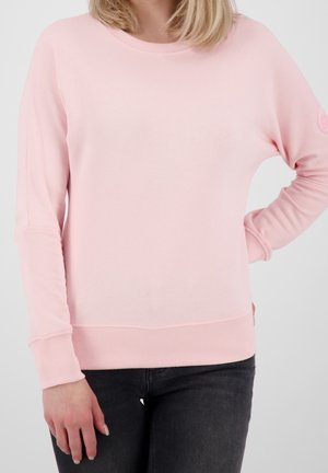 Kvinde iført lyserød langærmet sweater med ribbet manchetter og kant, kombineret med mørkegrå jeans, stående med den ene hånd på hoften.