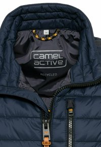 Marineblaue Daunenjacke mit gestepptem Muster, ausgestattet mit einem Reißverschluss, elastischem Kragen und "camel active"-Logo. Innen etikettiert als recyceltes Material.