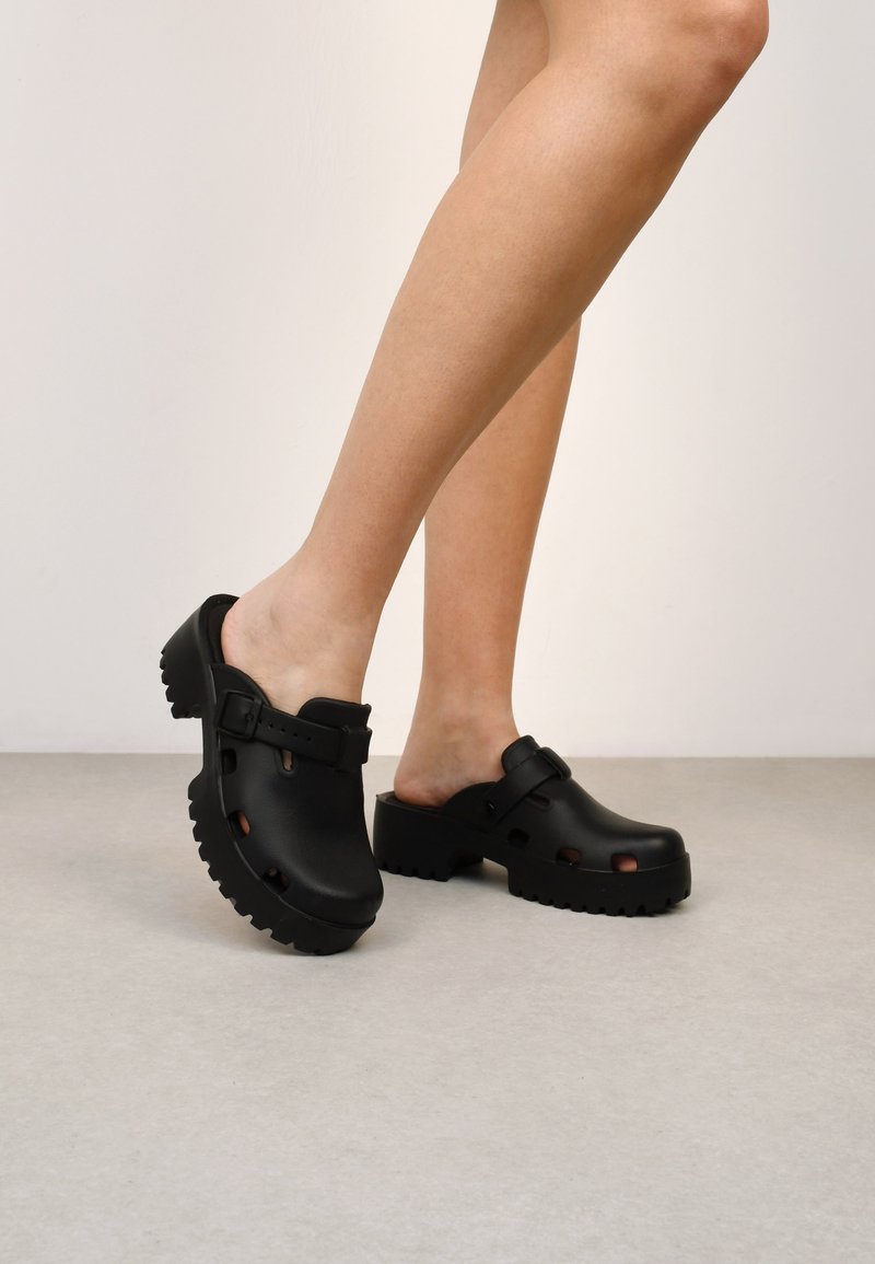 Zoccoli neri con superficie strutturata, dotati di cinturino con fibbia, apertura sul retro e fori ritagliati. Suola spessa e sagomata per aderenza e comfort.