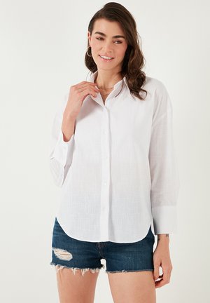 White button-up shirt with a textured surface, featuring long sleeves and a curved hem, paired with distressed denim shorts.