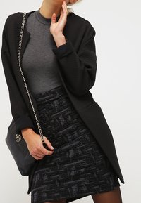Schwarze strukturierte Strickjacke mit offenem Vorderteil, getragen über einem grauen Top und einem gemusterten schwarzen Rock. Eine schwarze Handtasche mit einem silbernen Kettenriemen ist ebenfalls enthalten.