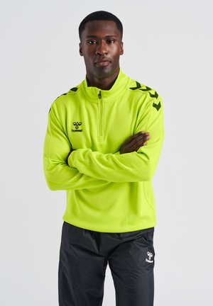 Mężczyzna w neonowo-żółtej sportowej kurtce z zamkiem do połowy i czarnych sportowych spodniach, z założonymi rękami, stoi na jednolitym tle.