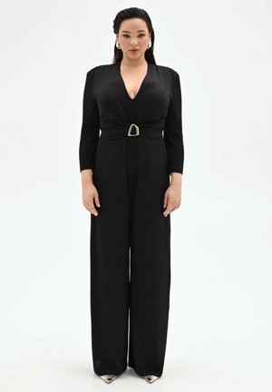 Donna in piedi di fronte, indossa una tuta lunga nera con scollo profondo e cintura con fibbia dorata, scarpe argentate con punta appuntita, orecchini dorati.