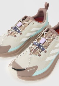 Beige en bruine sportschoenen met gemetste oppervlakken, GORE-TEX merk, reflecterende accenten en elastische veters, met een turquoise tussenzool.