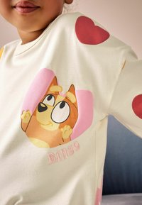 Camicetta a maniche lunghe di colore crema chiaro con un cane dei cartoni animati in rosa e colletto tondo, arricchita da grandi cuori rossi sulle maniche.