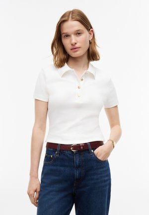 Jeune femme aux cheveux mi-longs portant un polo blanc à manches courtes, un jean bleu et une ceinture marron, la main dans la poche.