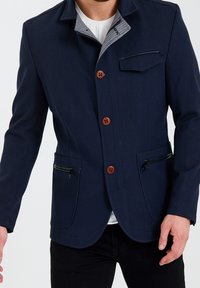 Marinblå jacka i strukturerad tyg, med bruna knappar, dragkedjefickor och en grå mönstrad krage. Skräddarsydd passform med en modern design.