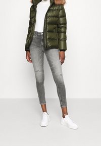 Olivgrön vadderad jacka med huva av fuskpäls, vit polotröja, gråa tajta jeans och vita sneakers. Texturerade material och figursydd design.