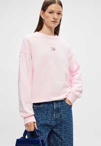 Jasno różowy sweter z luźnym krojem, okrągłym dekoltem, ściągaczami przy rękawach i małym logo. Połączony z ciemnoniebieskimi spodniami w deseń z denimu.