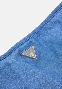 Zils denims audums ar teksturētu apdari, kurā ir trīsstūrveida brūns logo uzlīme ar "GUESS" un palmu.