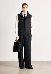 Μαύρο μπουφάν τύπου puffer με ψηλό γιακά, φορεμένο πάνω από ένα λευκό πουκάμισο και μαύρη γραβάτα. Συνοδεύεται από φαρδύ μαύρο παντελόνι και μαύρη δερμάτινη τσάντα.