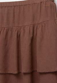 Gonna marrone a strati con vita arricciata, realizzata in tessuto leggero. Presenta una texture liscia e un sottile dettaglio del logo nella parte superiore.