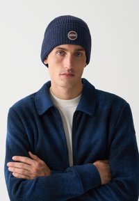 Bărbat îmbrăcat cu o căciulă tricotată de culoare albastră închisă cu logo-ul "COLMAR", o jachetă albastră închisă cu fermoar și un tricou alb, cu brațele încrucișate pe un fundal simplu.