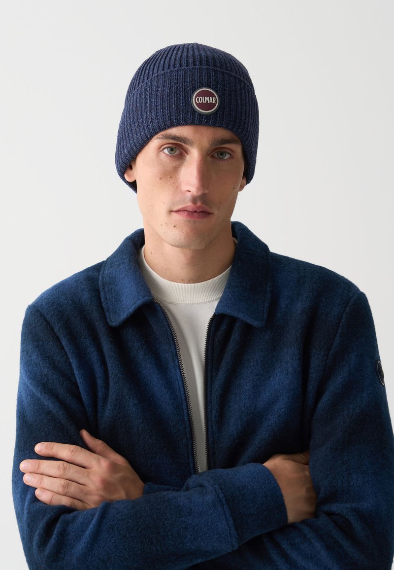 Bărbat îmbrăcat cu o căciulă tricotată de culoare albastră închisă cu logo-ul "COLMAR", o jachetă albastră închisă cu fermoar și un tricou alb, cu brațele încrucișate pe un fundal simplu.