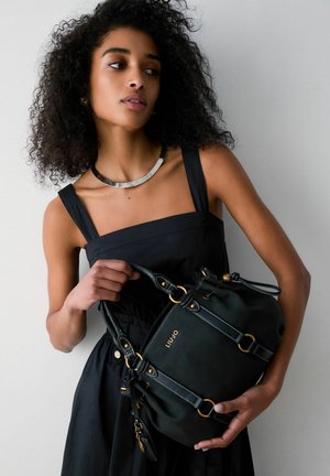 Woman with curly hair wearing a black dress and silver necklace, holding a black Liu Jo handbag with gold accents.