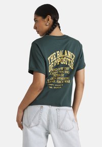 Green cotton t-shirt with short sleeves features yellow printed text on the back. Paired with light blue denim jeans.