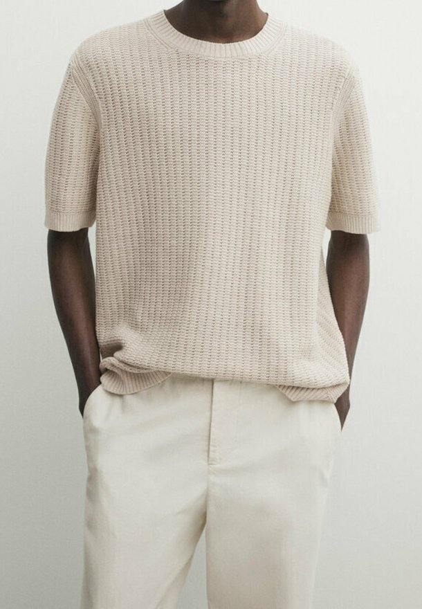 Beige knitted short-sleeve sweater with a textured pattern, round neckline, and relaxed fit, paired with light-colored trousers.