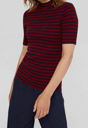 Kobieta ubrana w dopasowany, krótki rękaw, czerwono-czarny prążkowany golf i luźne, ciemnoniebieskie spodnie na jednolitym tle.