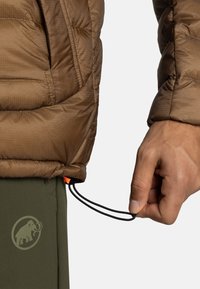 Brun oppustelig jakke med quiltet tekstur og justerbar bund, der har en orange toggle. En hånd holder fast i en sort snor nær grønne bukser.