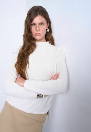 Ung kvinde med langt brunt hår, der krydser armene, iført hvid sweater, beige nederdel, sølvøreringe og armbånd, stående foran en ensfarvet baggrund.