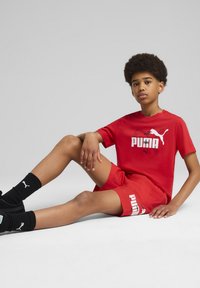 Conjunto de camiseta y pantalones cortos rojos de Puma, con un logo blanco. Calcetas y zapatos negros. Diseño ajustado, textura suave y estilo atlético.