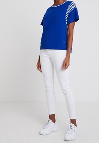 Blue short-sleeved t-shirt with striped shoulder accents, paired with white slim-fit jeans and white sneakers. Smooth cotton texture.