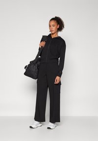 Μαύρη ζακέτα με φερμουάρ και φαρδύ παντελόνι cargo από μαλακό ύφασμα, συνδυασμένα με λευκά αθλητικά παπούτσια. Μια μαύρη τσάντα ώμου βρίσκεται στο πλάι της.