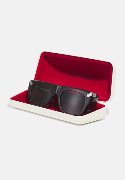 Sorte solbriller med en kraftig stel og mørke linser, placeret i et hvidt etui med en rød inderside, der har mærkets logo.