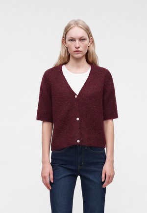 Femme portant un cardigan bordeaux à manches courtes en tissu duveteux avec des boutons, par-dessus une chemise blanche et un jean bleu foncé, debout devant un fond uni.